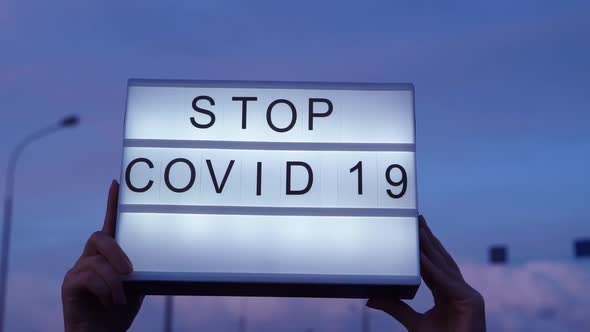 Hands Are Holding a Lightbox with a Sign Stop Covid-19, Pink Blue Sky on the Background. COVID-19