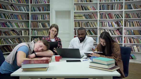 Guy which Sleeping on the Table and Three other Focused Multiracial Students Working on Laptop