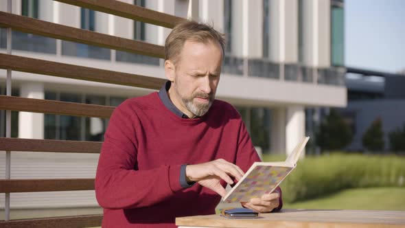 A Middleaged Handsome Caucasian Man Reads a Book As He Sits at a Table in a Park in an Urban Area