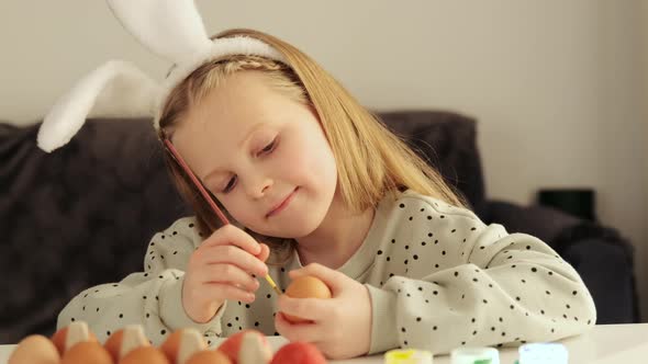 Little child girl with rabbit ears decorates Easter eggs with colored food paint.