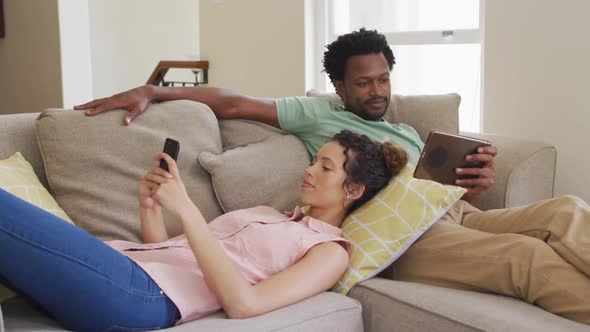 Happy biracial couple relaxing on sofa with smartphone and tablet