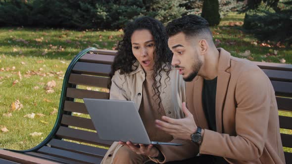 Excited Hispanic Couple Reading Good News on Laptop Sitting on Bench in Autumn Park Successful