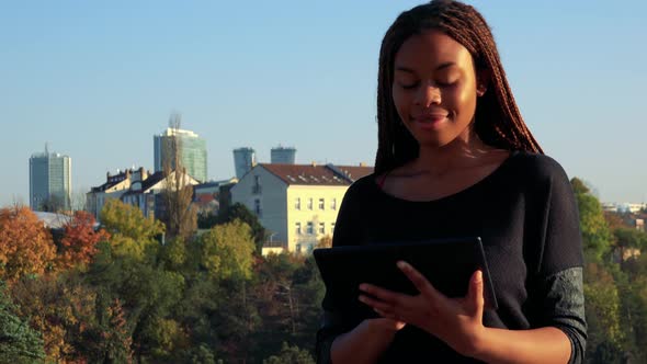 Young Black Woman Works on Tablet in Park - Buildings in the Background