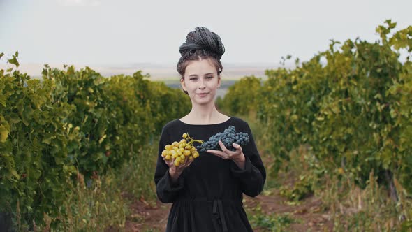 Young Smiling Woman with Dreadlocks Holding White and Black Grapes in Her Hands and Looks in the