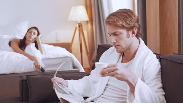 Young Concentrated Caucasian Male in White Bathrobe Drinking Coffee From Cup and Reading Morning