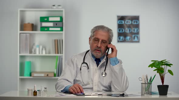 Aged Male Doctor Fills Out the Medical History and Advises Patient Using Mobile Phone About