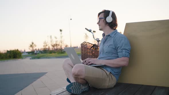 Freelancer Man Works at a Laptop in Headphones in a Park on a Bench at Sunset