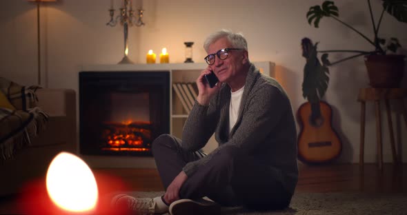 Happy Aged Caucasian Man Talking on Mobile Phone and Smiling Sitting Near Fireplace