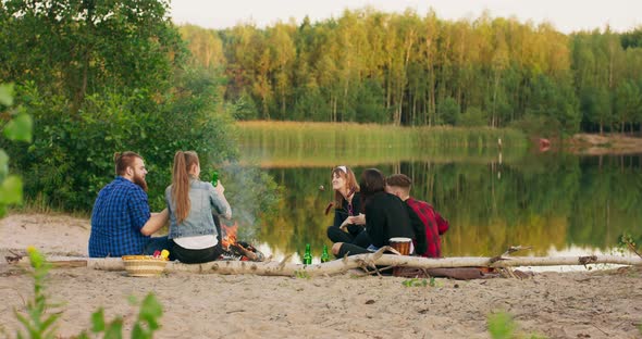 Group of Friends Having Party Around Bonfire on the Beach at Sunset Hipster Man with Beard Tells