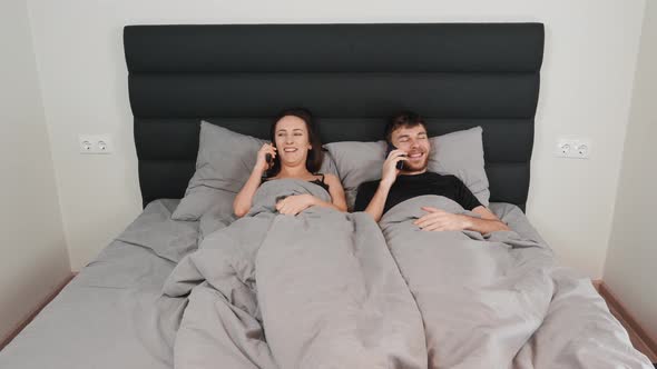 Happy couple lying in bed, talking on smartphones and laughing