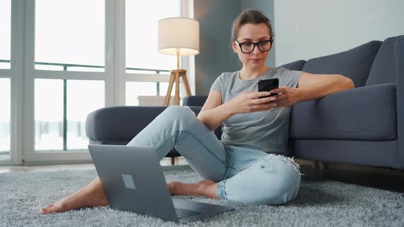 Casually Dressed Woman Sitting on Carpet with Laptop and Smartphone and Working in Cozy Room