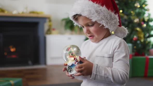 Surprised caucasian boy wearing santa hat playing with snow globe