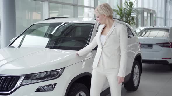 Portrait of Confident Blond Caucasian Woman Touching New Vehicle in Car Dealership and Crossing