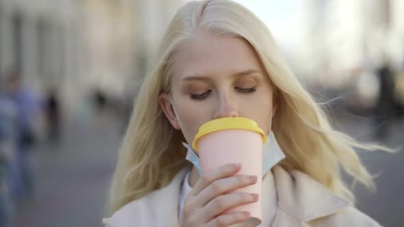 Young Blonde Woman Puts Down Her Medical Mask Drinks Coffee and Puts on the Mask Again in the Street