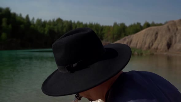 A Man with Glasses and a Hat Plays the Saxophone on a Summer Day on the Shore of a Lake, Behind