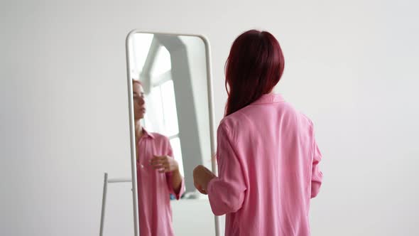 Young Woman in a Pink Shirt Straightens Her Red Hair Near the Mirror