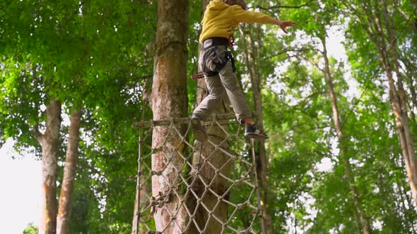 Slowmotion Shot of a Little Boy in a Safety Harness Climbs on a Zipline in Treetops in a Forest