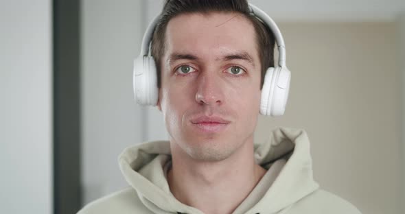 Portrait of a Stylish Man in White Headphones Listening to Music and Looking at the Camera Indoors