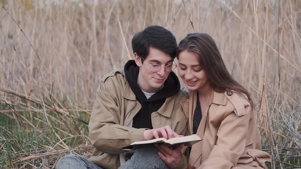 Young Couple Sitting in Field and Reading Book Aloud