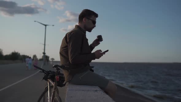Man Cyclist Resting Overlooking Sea Drinking Coffee and Using Smartphone While Sitting on Embankment