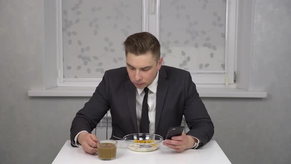 A Young Man in a Suit Eats Cereal and Drinks Coffee and Uses a Smartphone