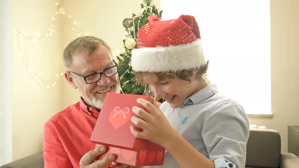 Grandpa Wearing a Santa Hat Makes a Christmas Present To His Teenage Grandson. The Boy Is Delighted