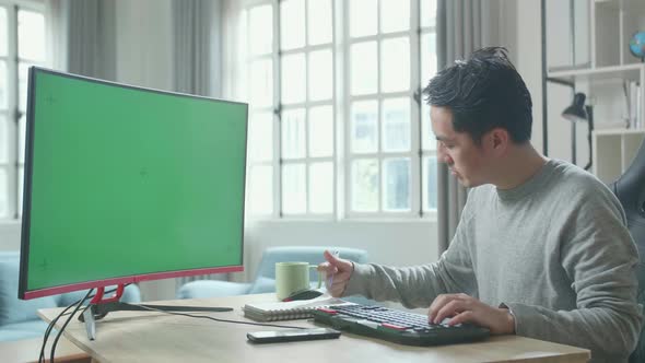 Asian Man Looking At Green Screen Desktop Computer And Writing In Notebook While Working At Home
