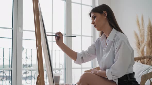 Young Woman Draws Painting Sitting on Chair in Art Studio