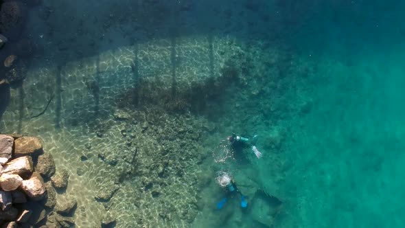 A high view looking down at 2 scuba divers swimming in clear water next to a rocky outcrop