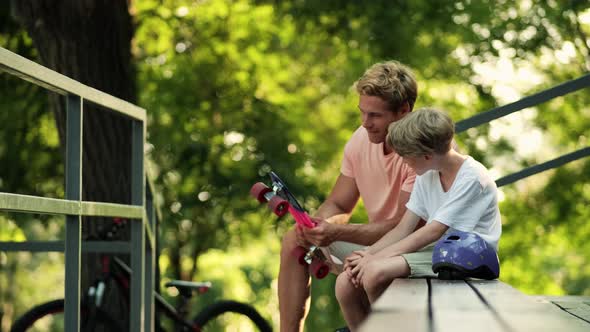 A smiling father is talking with his little boy while holding a penny board sitting on the bench