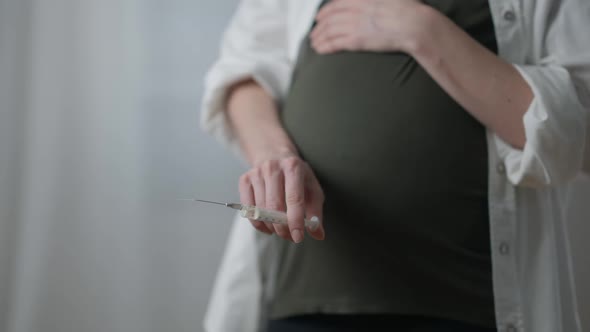 Unrecognizable Pregnant Addicted Woman with Syringe Standing Indoors Touching Belly