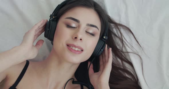 Happy Girl Listening to Music on Bed Flirts at Camera Twists Headphones' Wire