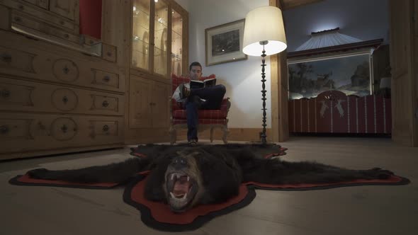 A man reads a book in a chair with a bearskin rug at a ski resort.