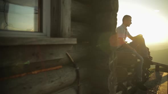 Young Man Sit Near Old Wooden House. Sun Beams. Slow Motion