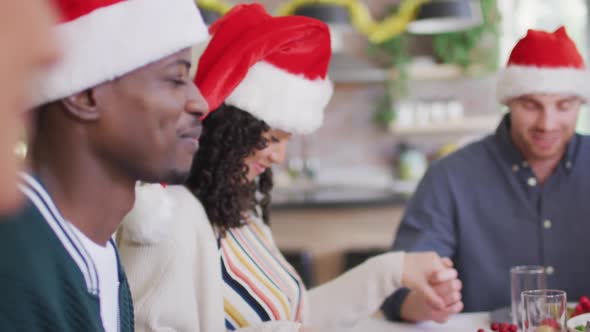 Happy group of diverse friends in santa hats praying, celebrating meal at christmas time