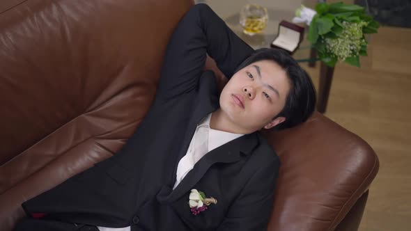 Top View of Sad Asian Groom Lying on Comfortable Couch Thinking with Blurred Ring Box Bouquet and
