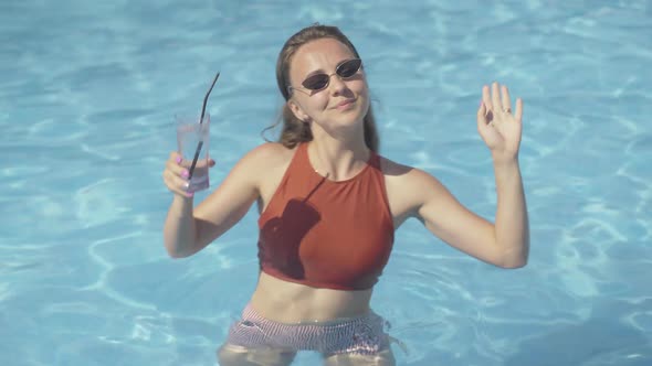 Middle Shot of Positive Young Woman Drinking Cocktail and Dancing in Water in Pool. Portrait of