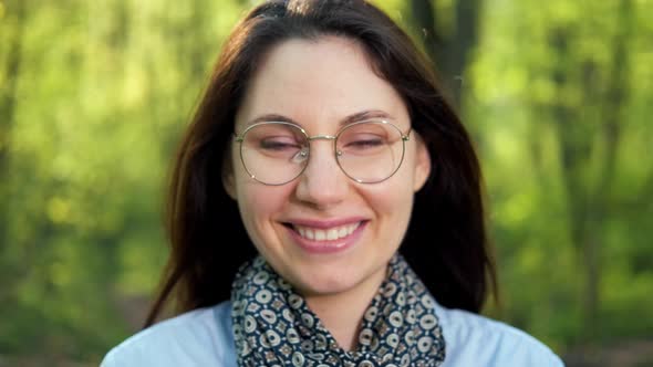 A Woman in Glasses and a Scarf Looks at the Camera and Smiles in the Park