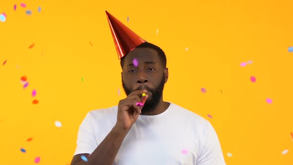 Upset Afro-American Man in Party Hat Whistling in Horn Under Falling Confetti