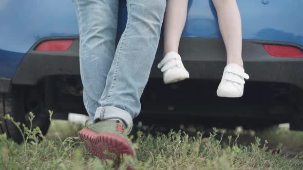 Legs of Little Girl and Young Man Sitting on Car Trunk Outdoors