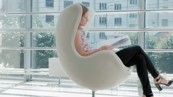 Attractive Businesswoman Sitting on a White Chair in a Glass Office and Checks Documents