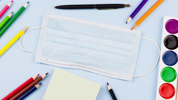 School Supplies Lie on a Pastel Blue Background with a Medical Mask