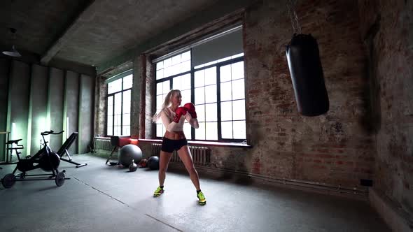 Blonde Slender Female Boxer in Shorts and Top, Fighting with a Punching Bag in the Gym in Gloves