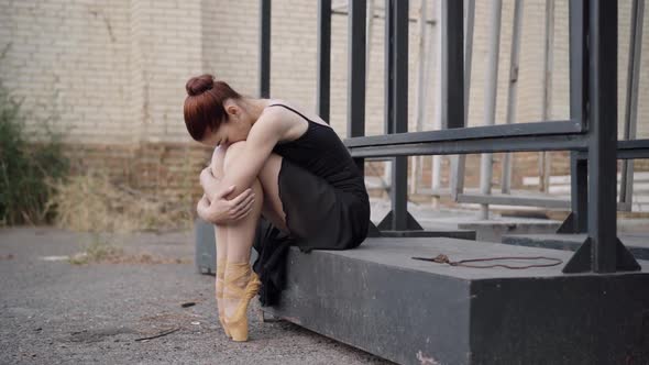 Sad Slim Caucasian Ballerina in Black Dress Sitting Outdoors Hugging Knees