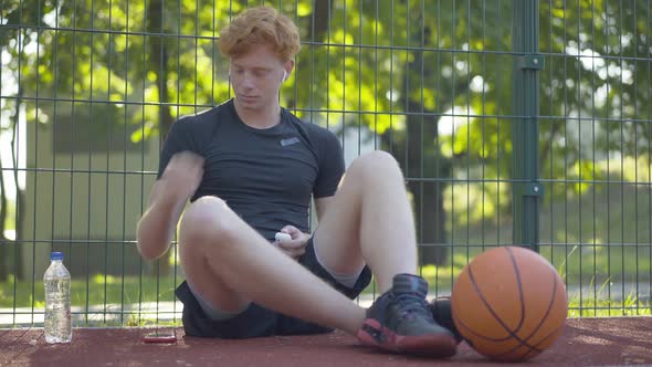 Wide Shot Portrait of Satisfied Basketball Player Turning on Music in Headphones. Confident Redhead