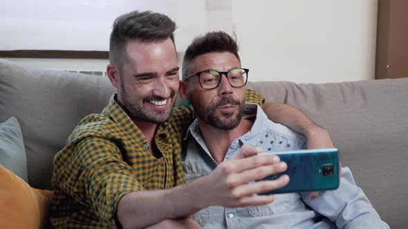 Gay Couple Sitting in the Sofa At Home Making Video Call On Mobile Phone