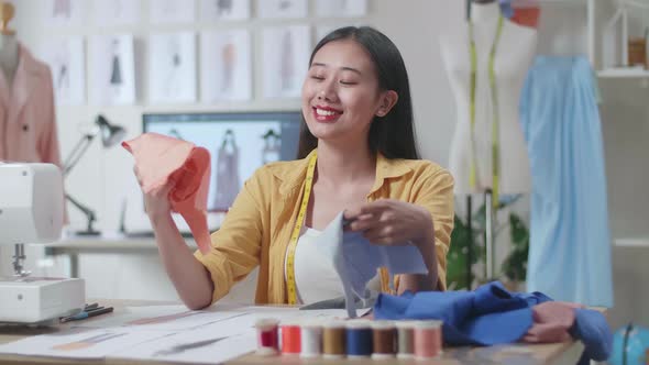 Asian Female Designer With Sewing Machine Holding And Looking At The Fabrics While Designing Clothes