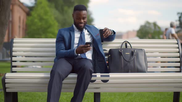 Young African Businessman Using Mobile Phone and Holding Cup of Takeout Coffee on Bench Outside