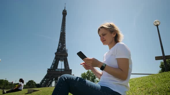 Woman is Taking Selfie Using Smartphone Sitting Near Eiffel Tower in Paris in Daytime Wide Shot