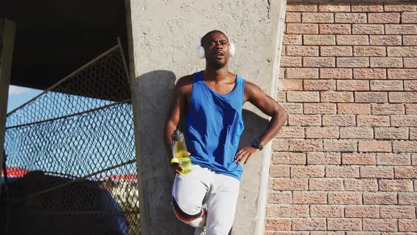 African american man wearing headphones drinking from water bottle taking break in exercise outdoors